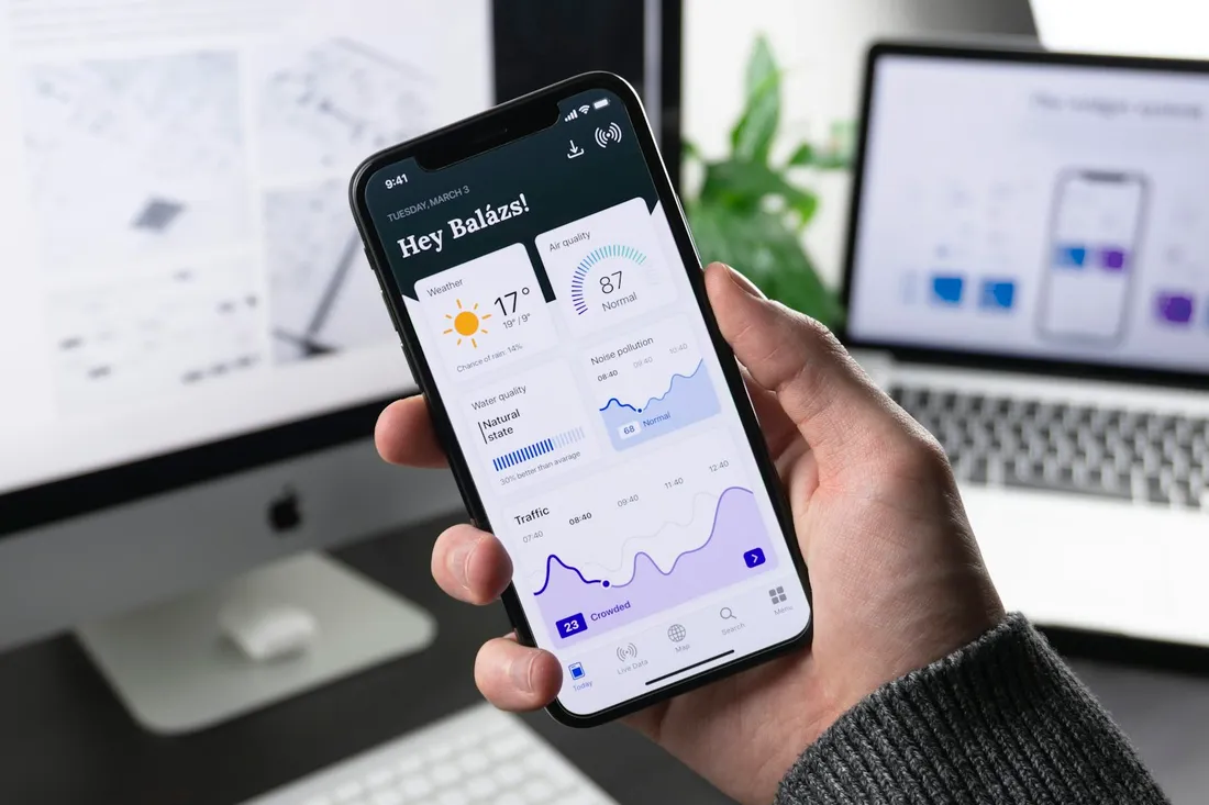 Hand holding a phone with an app open in front of a desktop design workspace